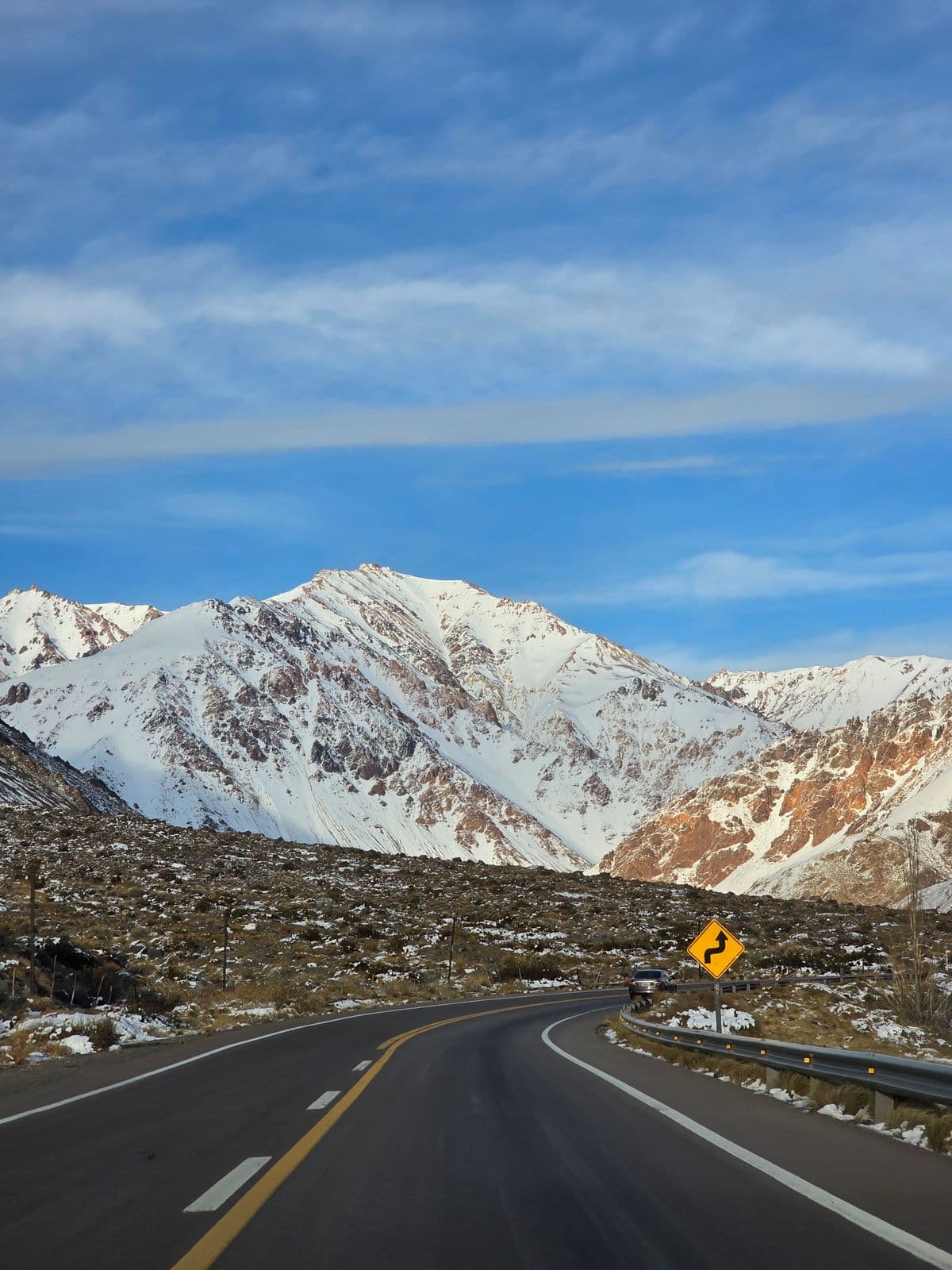 Ruta de montaña con picos nevados y un cartel de curva al costado